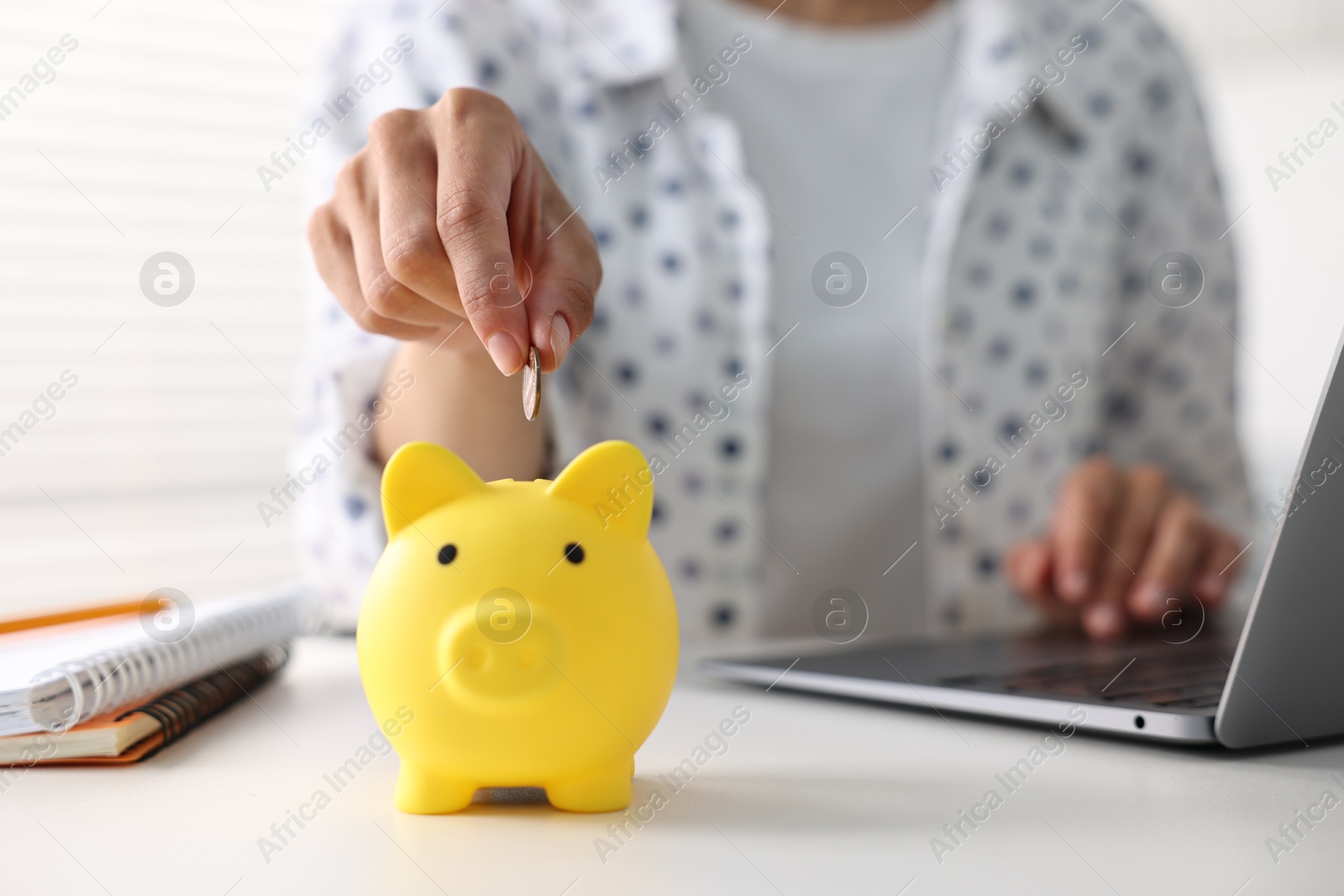 Photo of Woman putting coin into piggy bank at white table indoors, closeup