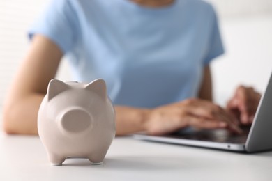 Photo of Woman using laptop at white table indoors, focus on piggy bank