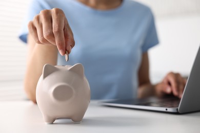 Photo of Woman putting coin into piggy bank at white table indoors, closeup