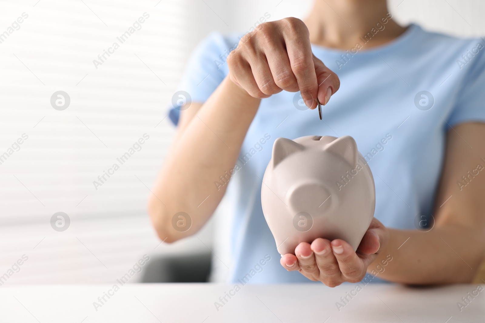 Photo of Woman putting coin into piggy bank at white table indoors, closeup