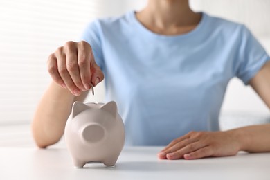 Photo of Woman putting coin into piggy bank at white table indoors, closeup