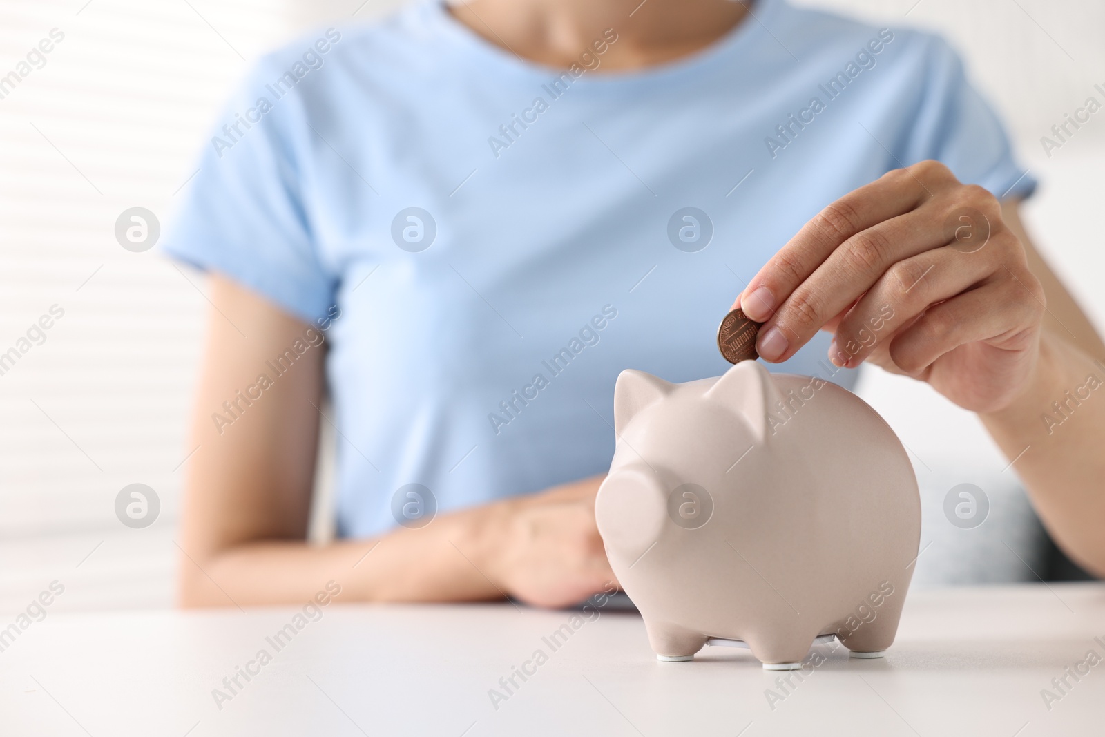 Photo of Woman putting coin into piggy bank at white table indoors, closeup