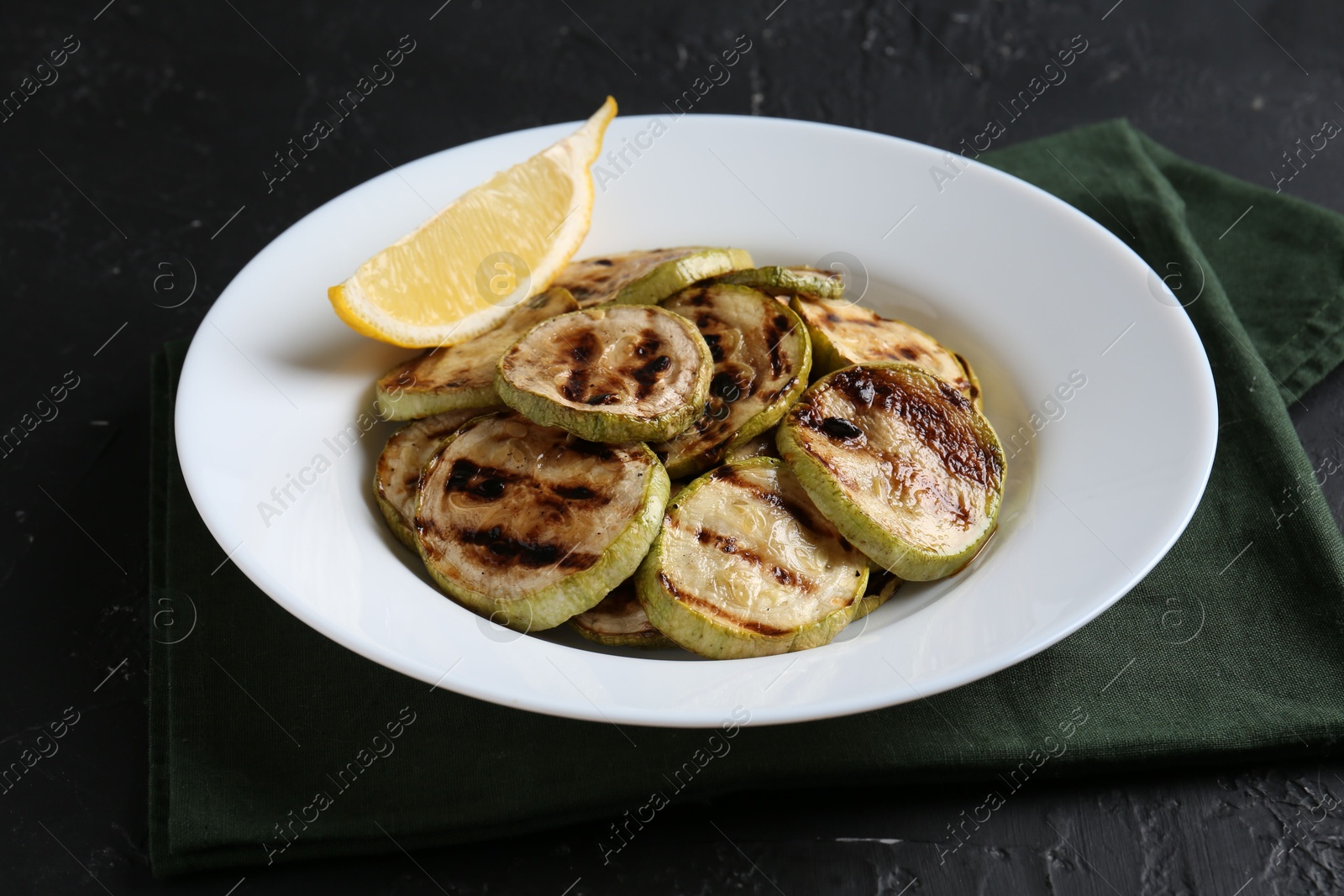 Photo of Tasty grilled zucchini slices with lemon on black textured table