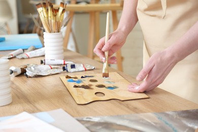 Photo of Woman with brush using palette at wooden table in drawing studio, closeup