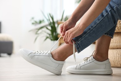 Photo of Woman tying shoelace of white sneaker indoors, closeup