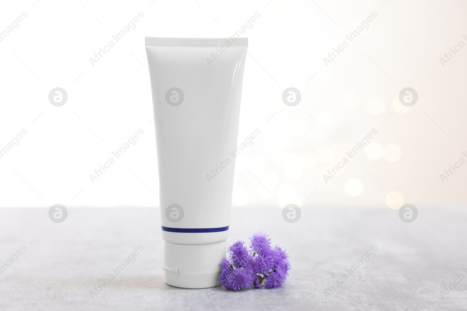 Photo of Tube of cosmetic product and ageratum flowers on gray table against light background with blurred lights