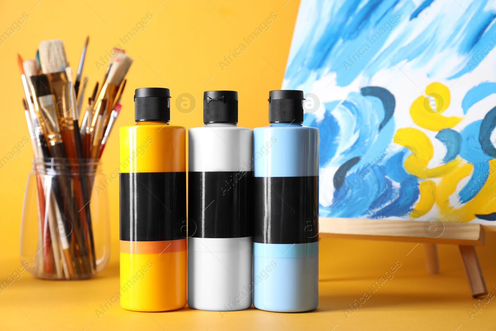 Photo of Acrylic paints of different colors, small easel with abstract picture and brushes on orange background