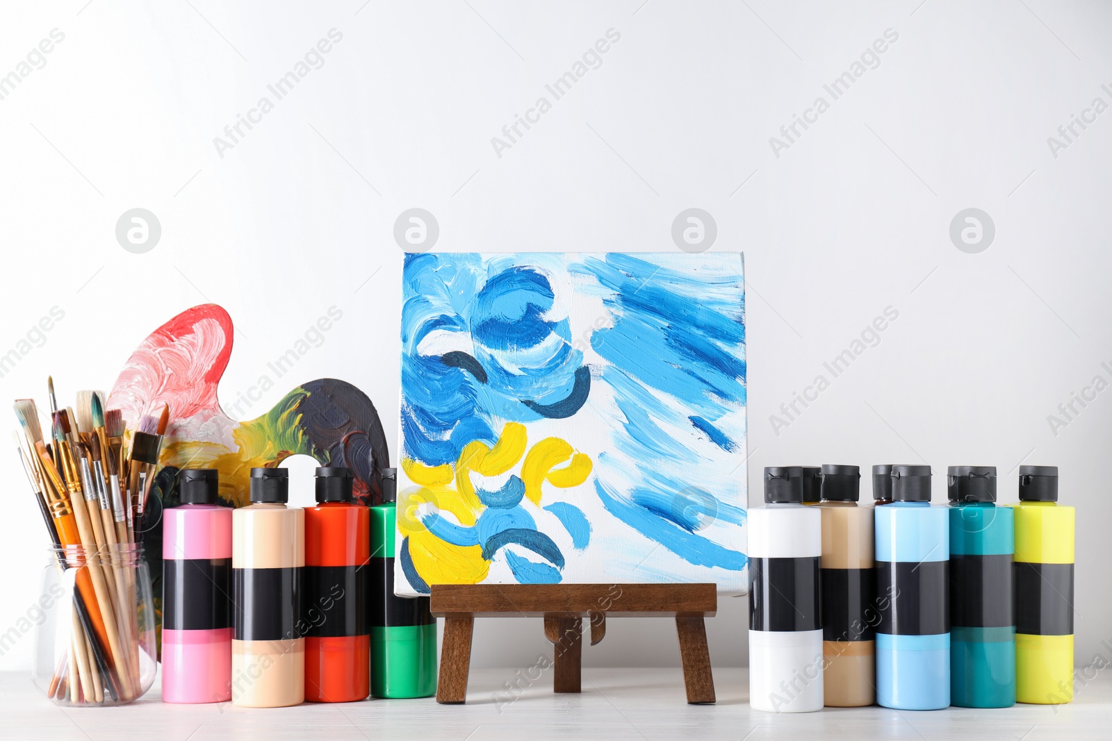 Photo of Acrylic paints of different colors, small easel with abstract picture, palette and brushes on wooden table