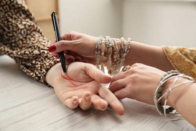 Fortune teller reading lines on woman's palm at white wooden table, closeup. Chiromancy