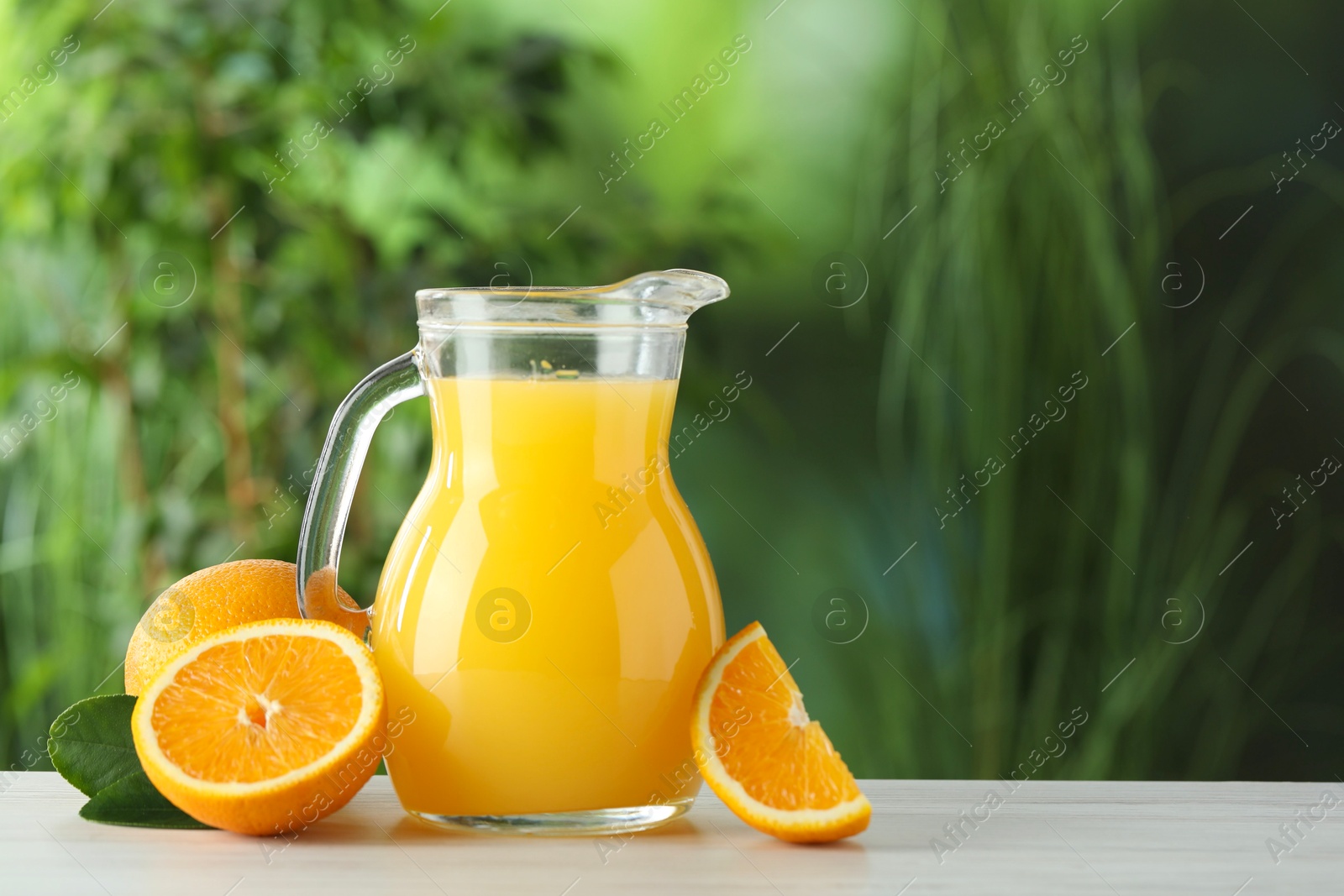 Photo of Tasty orange juice in glass jug, fresh fruits and green leaves on white wooden table outdoors, space for text