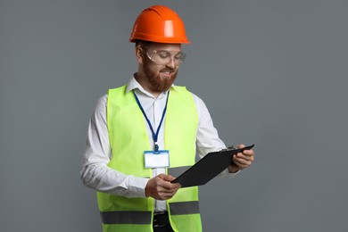 Photo of Engineer in hard hat and goggles holding clipboard on grey background