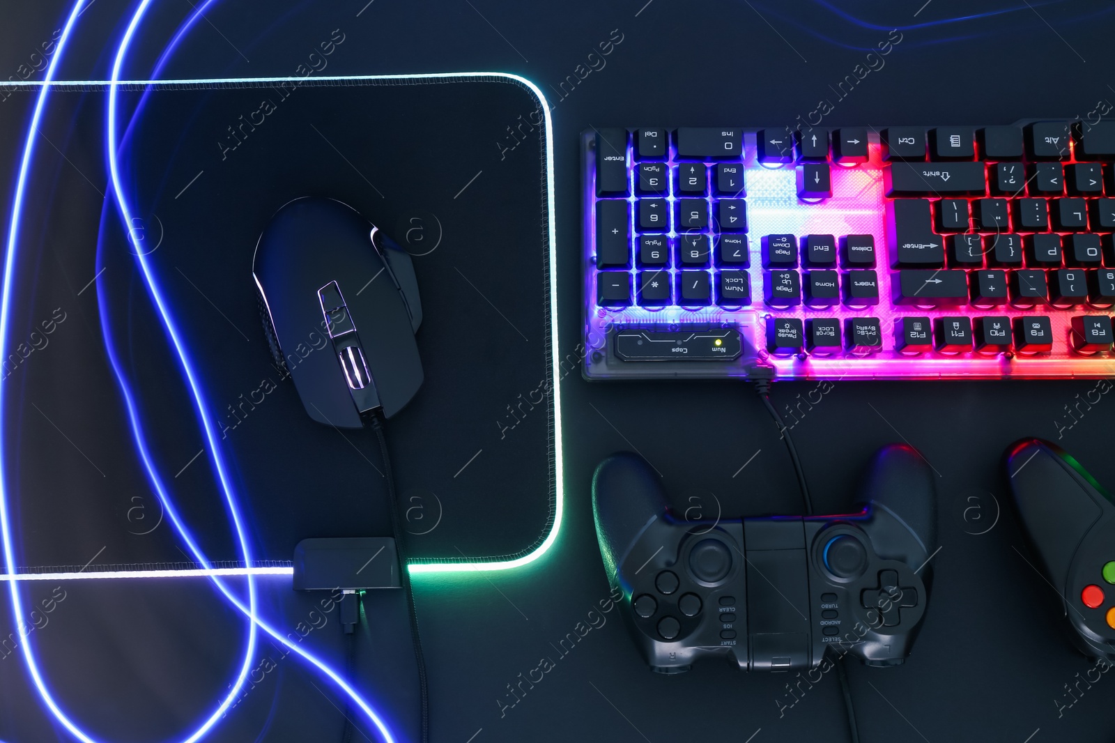 Photo of Computer mouse, RGB keyboard and game controllers in neon lights on black background, flat lay