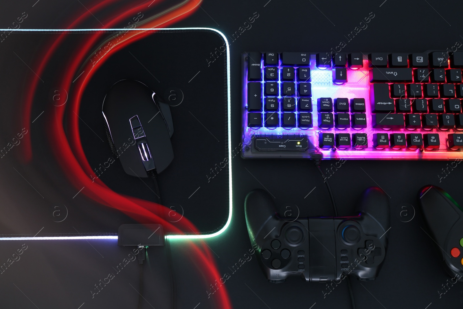 Photo of Computer mouse, RGB keyboard and game controllers in neon lights on black background, flat lay