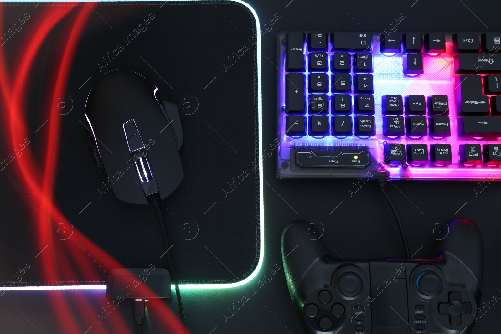 Photo of Computer mouse, RGB keyboard and game controller in neon lights on black background, flat lay