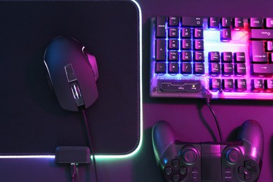 Computer mouse, RGB keyboard and game controller in neon lights on dark background, flat lay