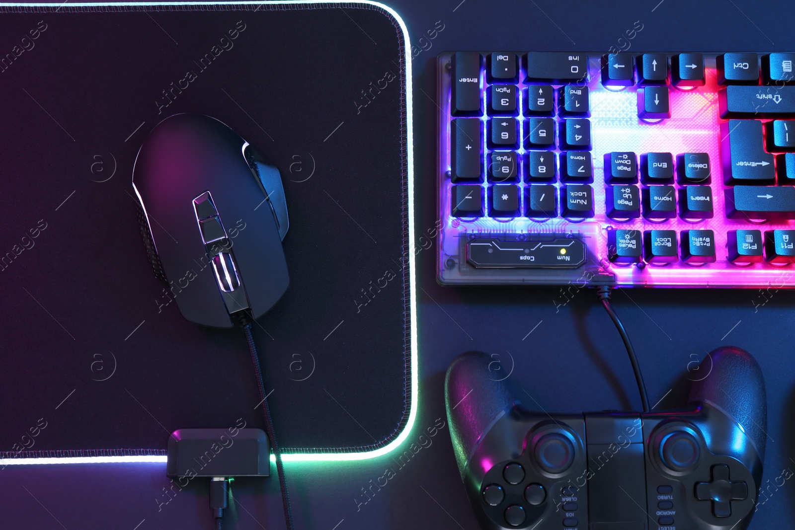 Photo of Computer mouse, RGB keyboard and game controller in neon lights on dark background, flat lay