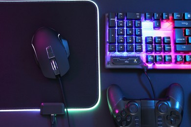 Photo of Computer mouse, RGB keyboard and game controller in neon lights on dark background, flat lay