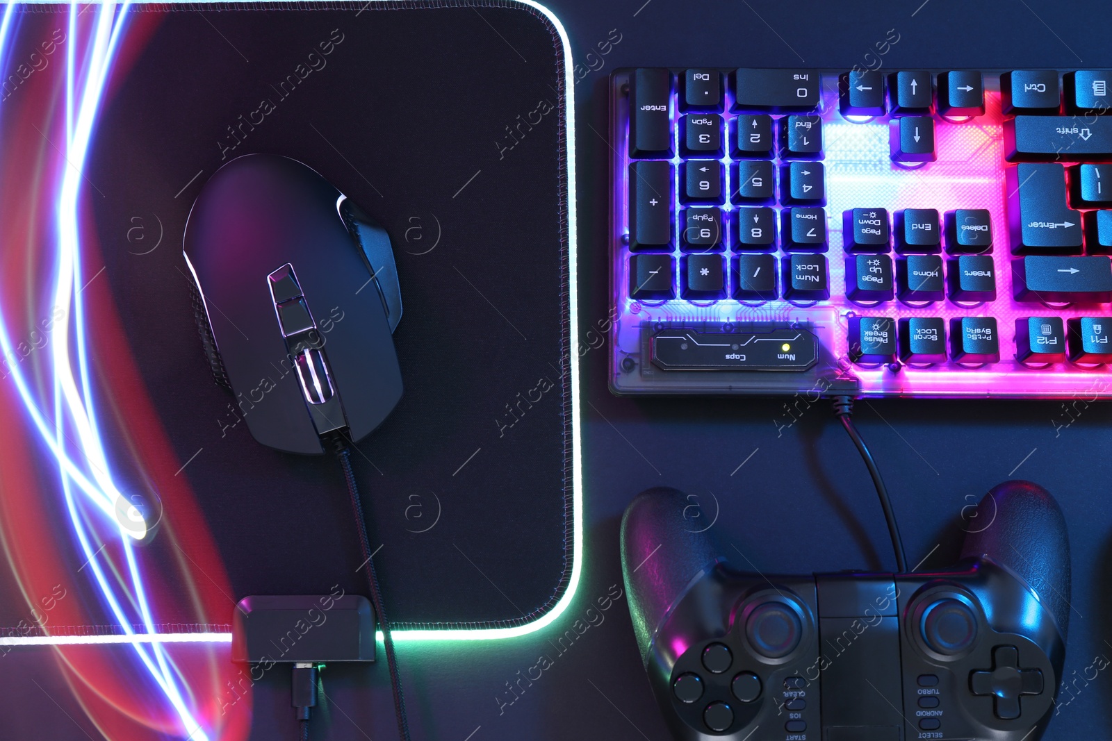 Photo of Computer mouse, RGB keyboard and game controller in neon lights on dark background, flat lay