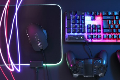 Photo of Computer mouse, RGB keyboard and game controller in neon lights on dark background, flat lay