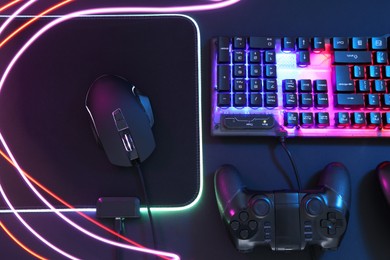 Photo of Computer mouse, RGB keyboard and game controller in neon lights on dark background, flat lay