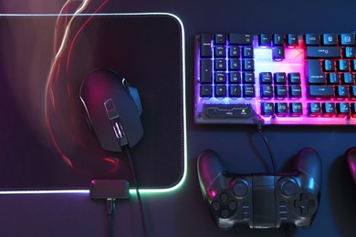 Photo of Computer mouse, RGB keyboard and game controller in neon lights on dark background, flat lay