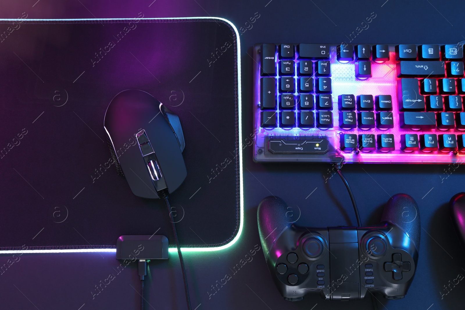 Photo of Computer mouse, RGB keyboard and game controller in neon lights on dark background, flat lay