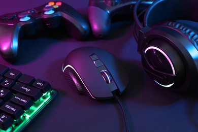 Photo of Computer mouse, headset, RGB keyboard and game controllers in neon lights on dark background, closeup