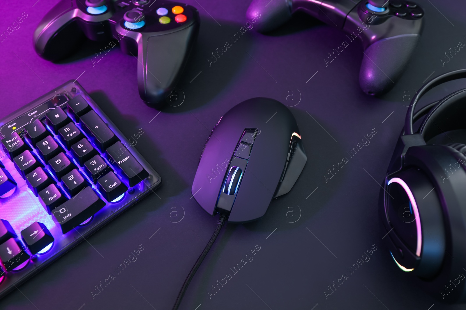 Photo of Computer mouse, headset, RGB keyboard and game controllers in neon lights on dark background, closeup