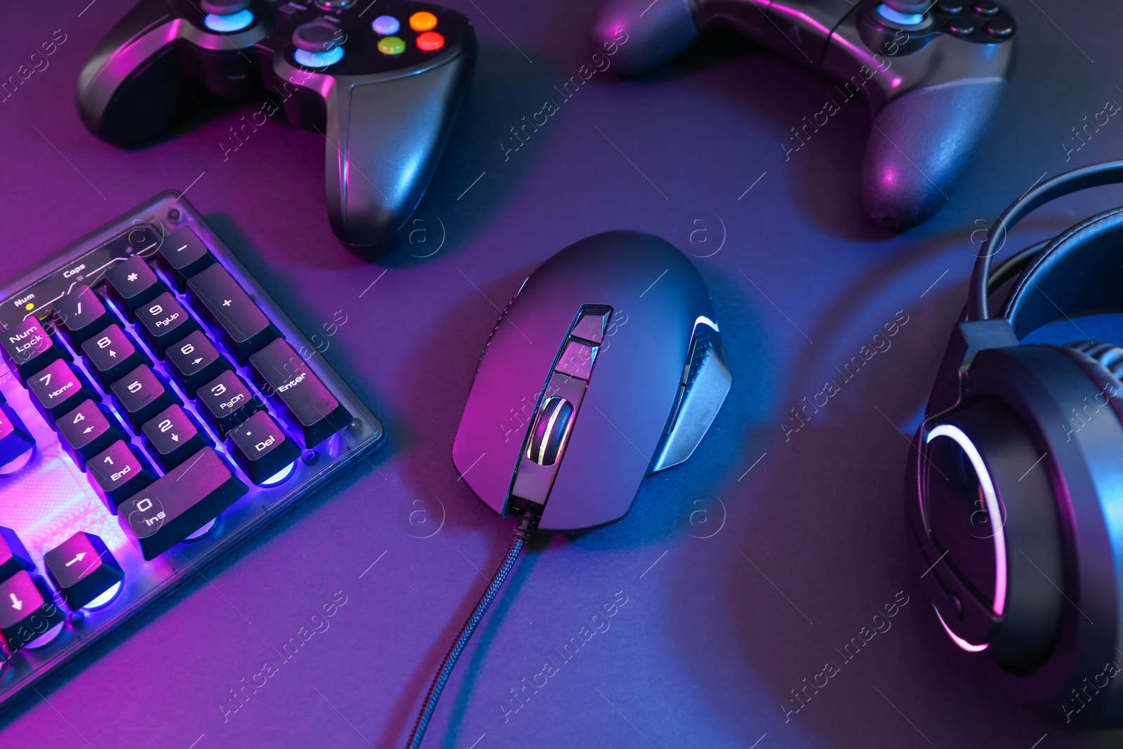 Photo of Computer mouse, headset, RGB keyboard and game controllers in neon lights on dark background, closeup