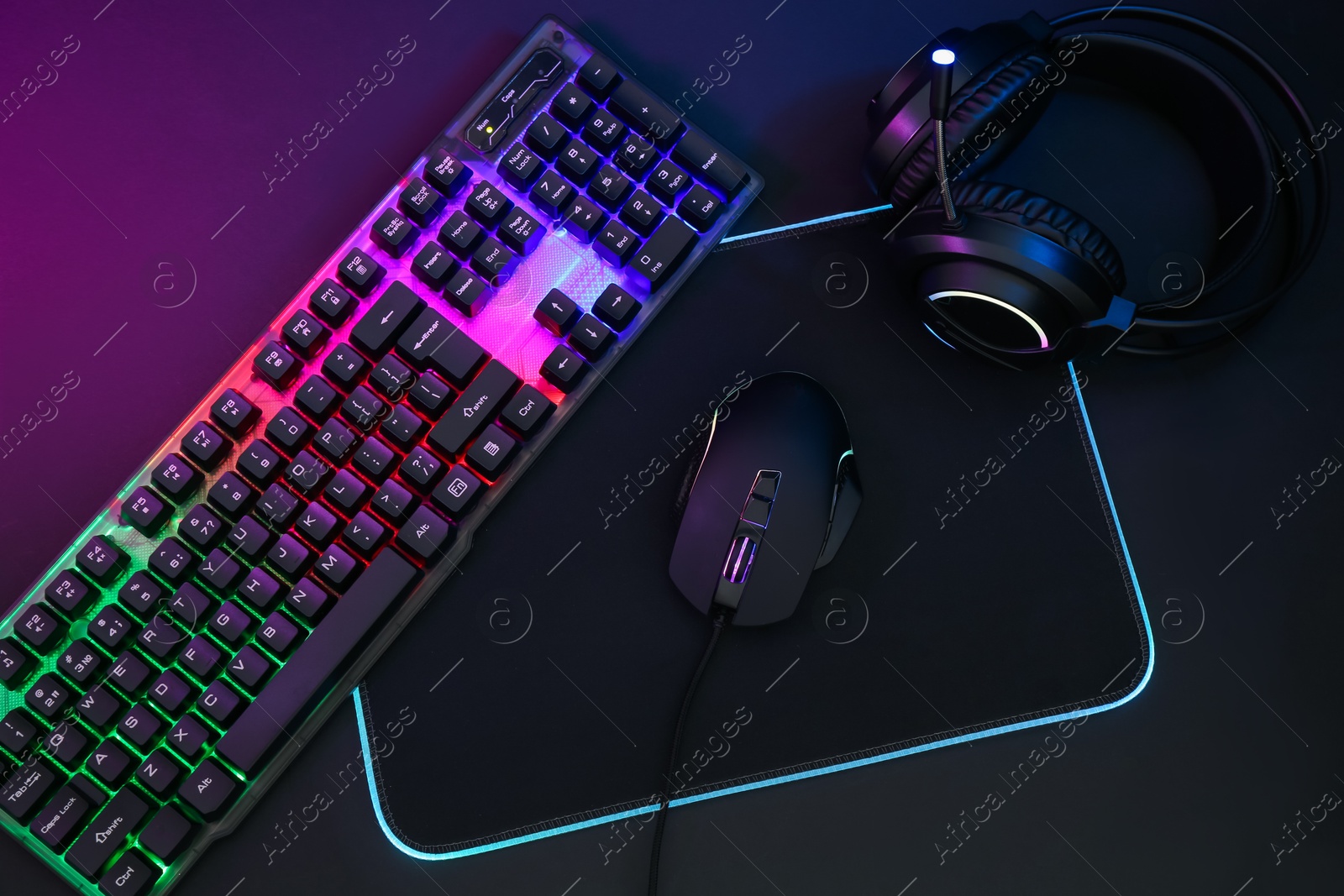 Photo of Computer mouse, headset and RGB keyboard in neon lights on dark background, flat lay