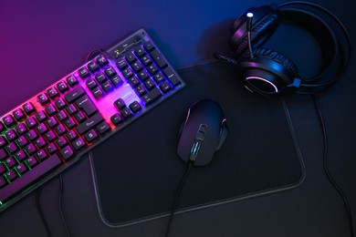 Computer mouse, headset and RGB keyboard in neon lights on dark background, flat lay