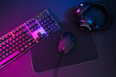 Computer mouse, headset and RGB keyboard in neon lights on dark background, flat lay