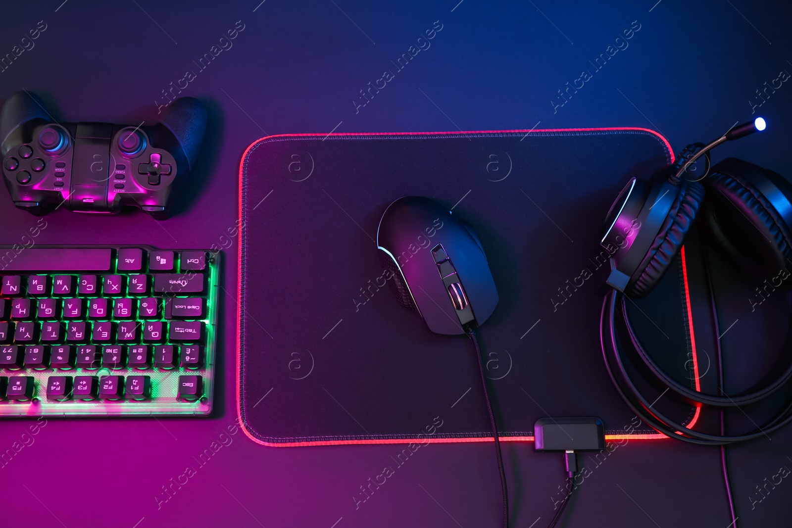 Photo of Computer mouse, headset, RGB keyboard and game controller in neon lights on dark background, flat lay