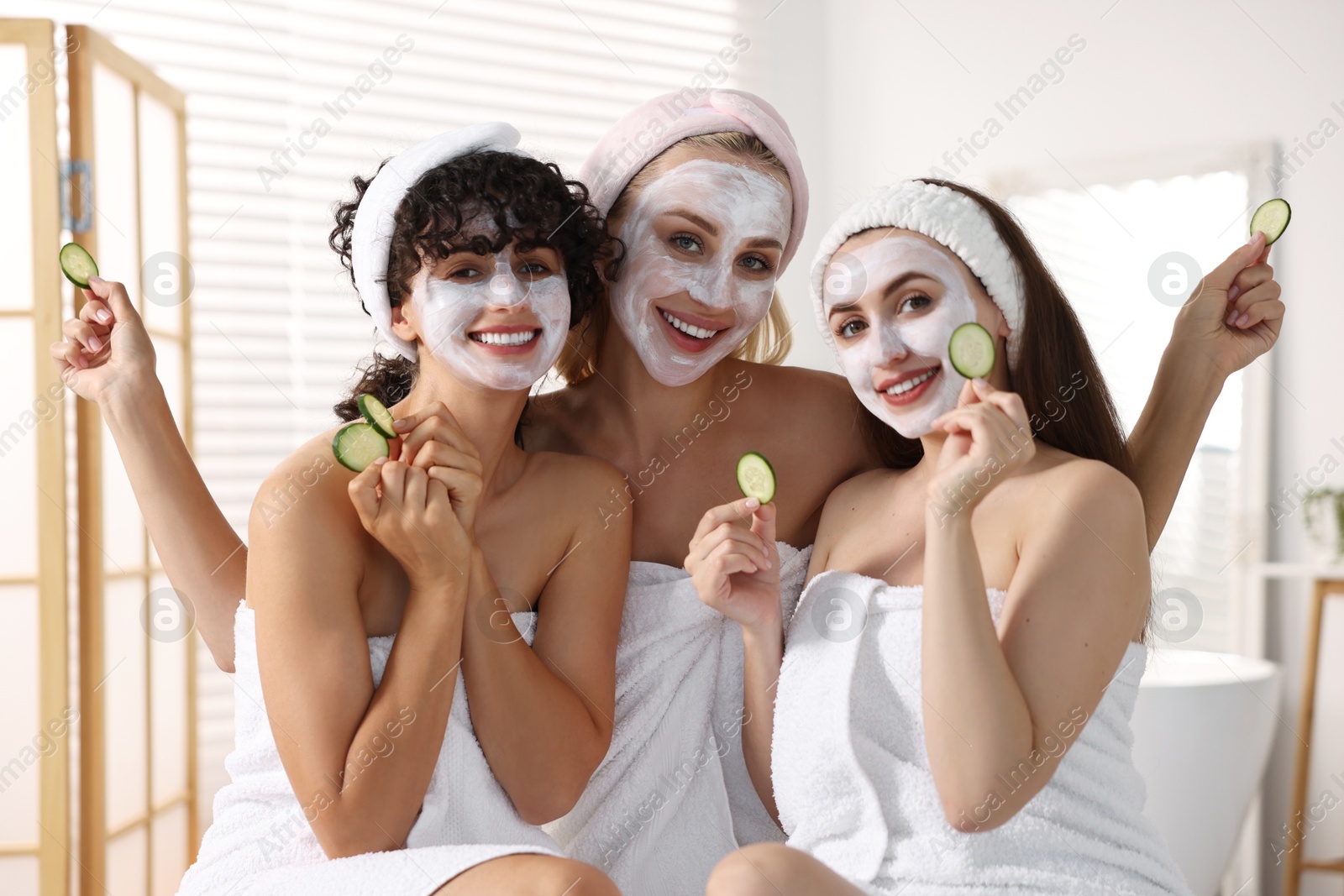 Photo of Happy friends with facial masks and cucumber slices in bathroom. Spa party