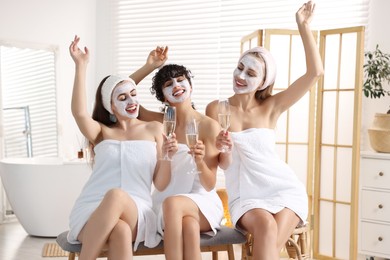 Photo of Happy friends with facial masks and glasses of sparkling wine in bathroom. Spa party