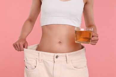 Photo of Weight loss concept. Woman with cup of diet tea and big pants on pink background, closeup