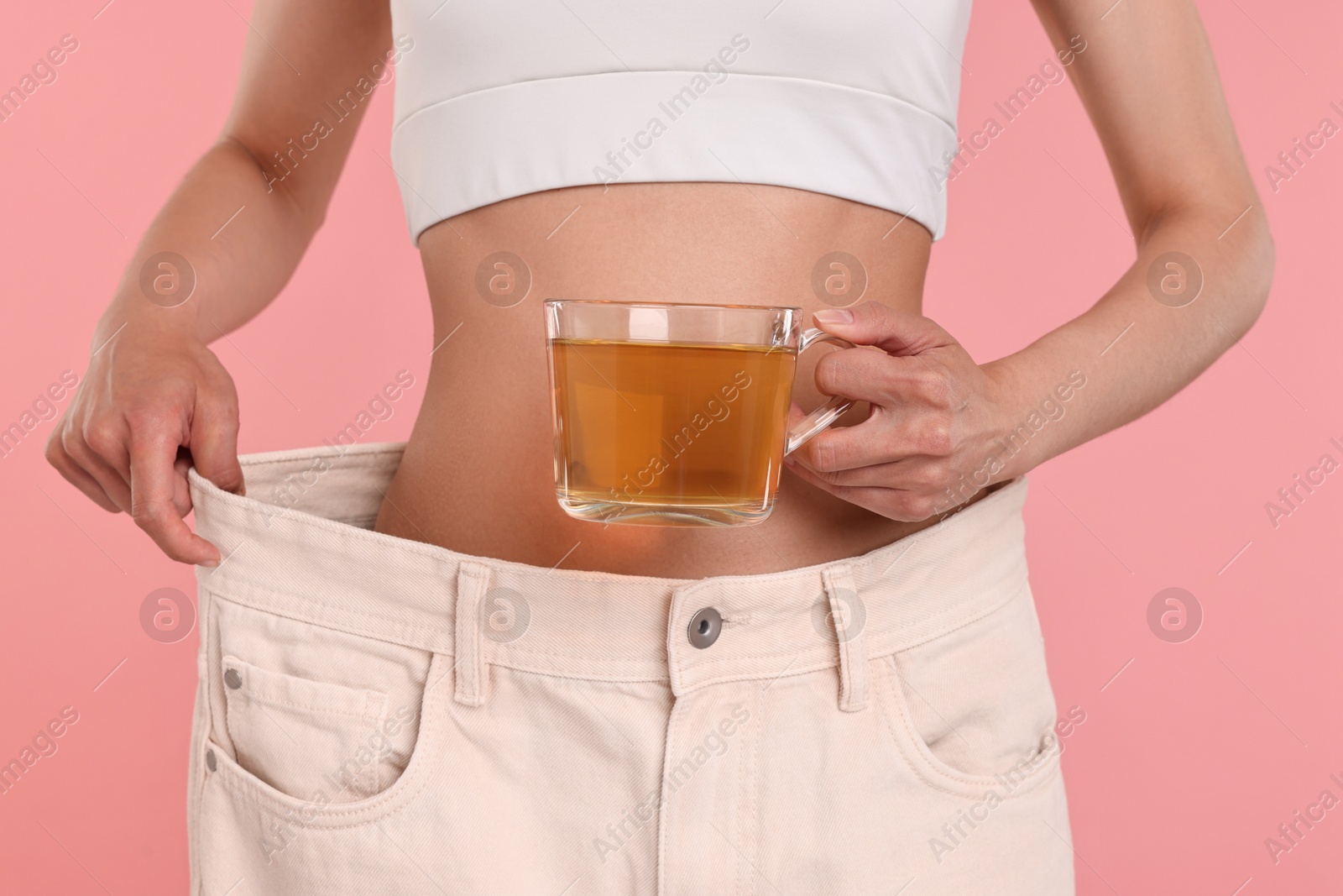 Photo of Weight loss concept. Woman with cup of diet tea and big pants on pink background, closeup