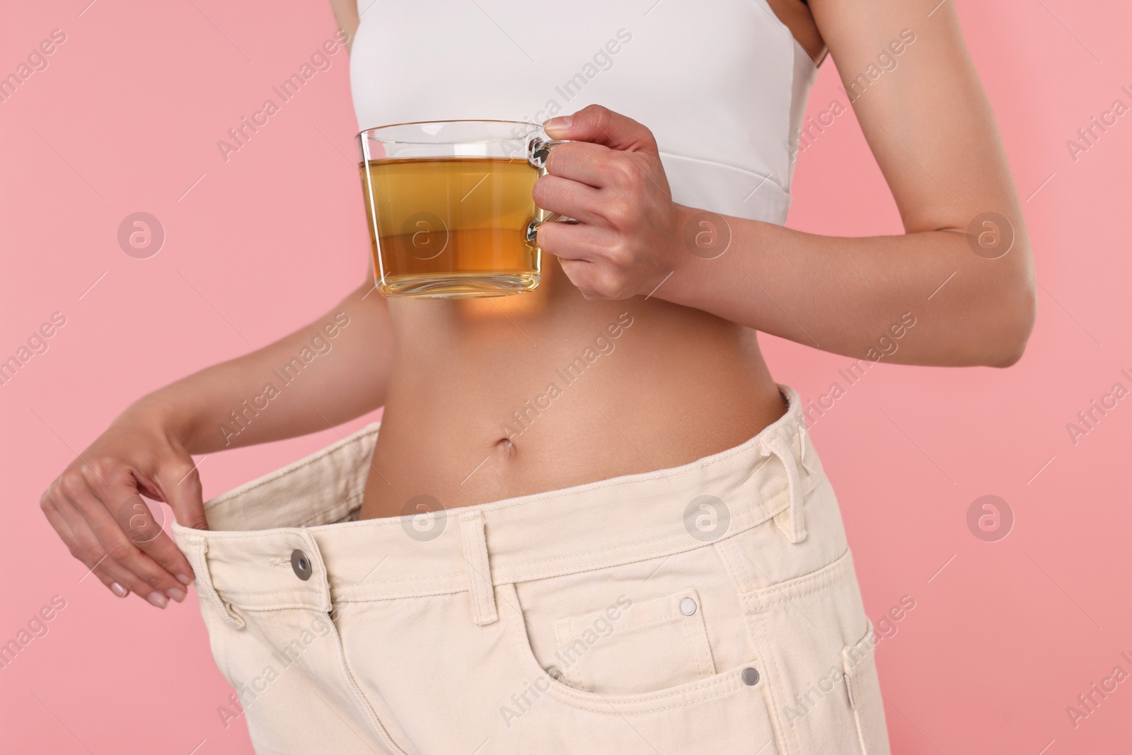 Photo of Weight loss concept. Woman with cup of diet tea and big pants on pink background, closeup