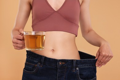 Photo of Weight loss concept. Woman with cup of diet tea and big pants on beige background, closeup