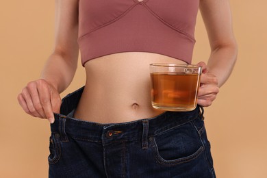 Photo of Weight loss concept. Woman with cup of diet tea and big pants on beige background, closeup