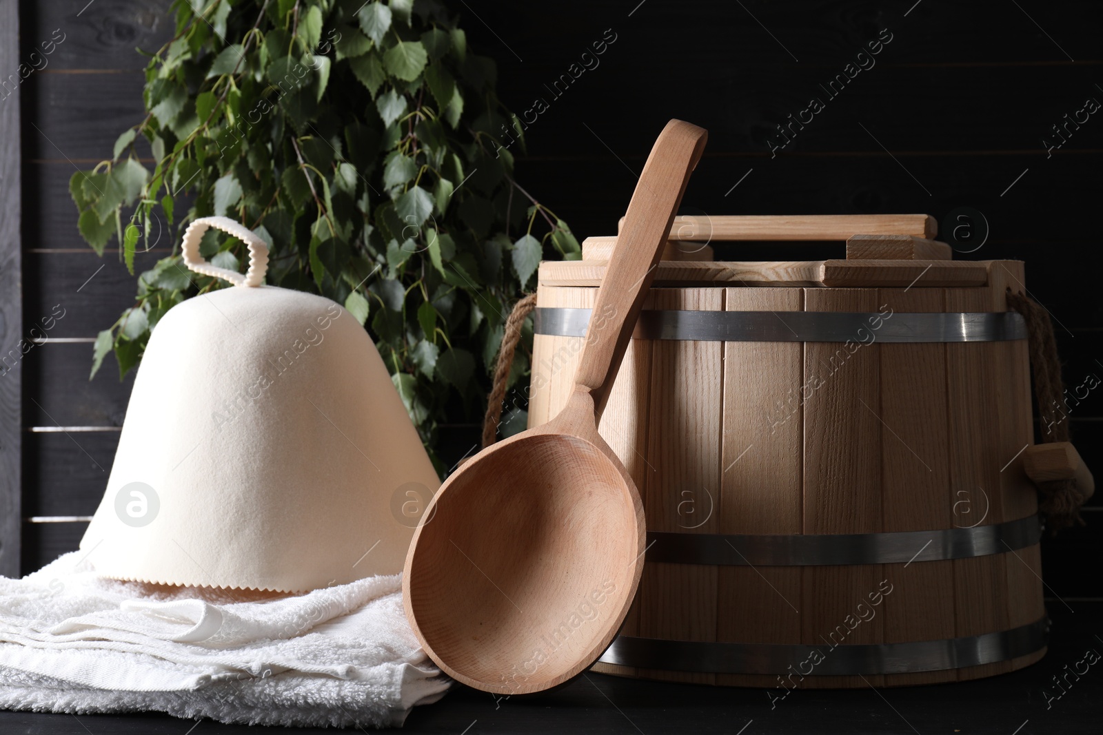 Photo of Sauna equipment. Wooden bucket, towel, felt wool hat and ladle on black surface