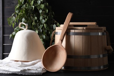 Photo of Sauna equipment. Wooden bucket, towel, felt wool hat and ladle on black surface