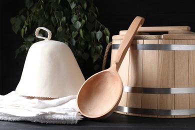 Photo of Sauna equipment. Wooden bucket, towel, felt wool hat and ladle on black surface