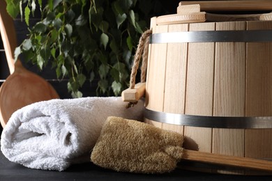 Photo of Sauna equipment. Wooden bucket, towel and loofah on black table