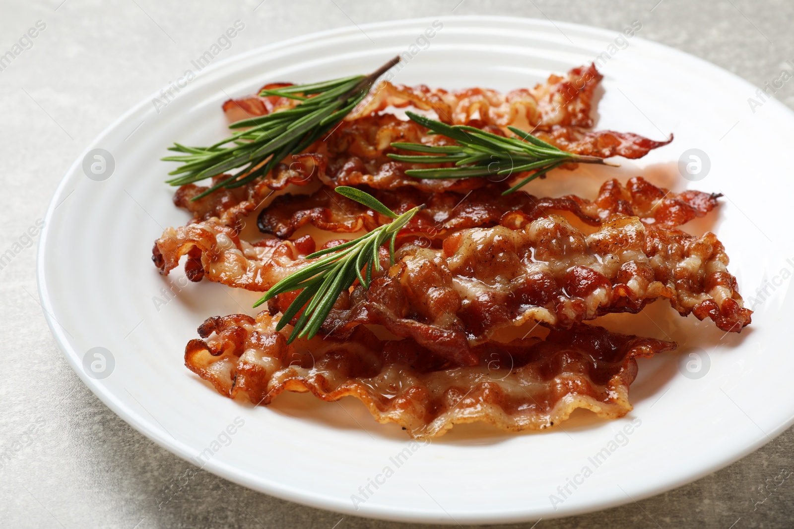 Photo of Slices of tasty fried bacon and rosemary on light grey table, closeup