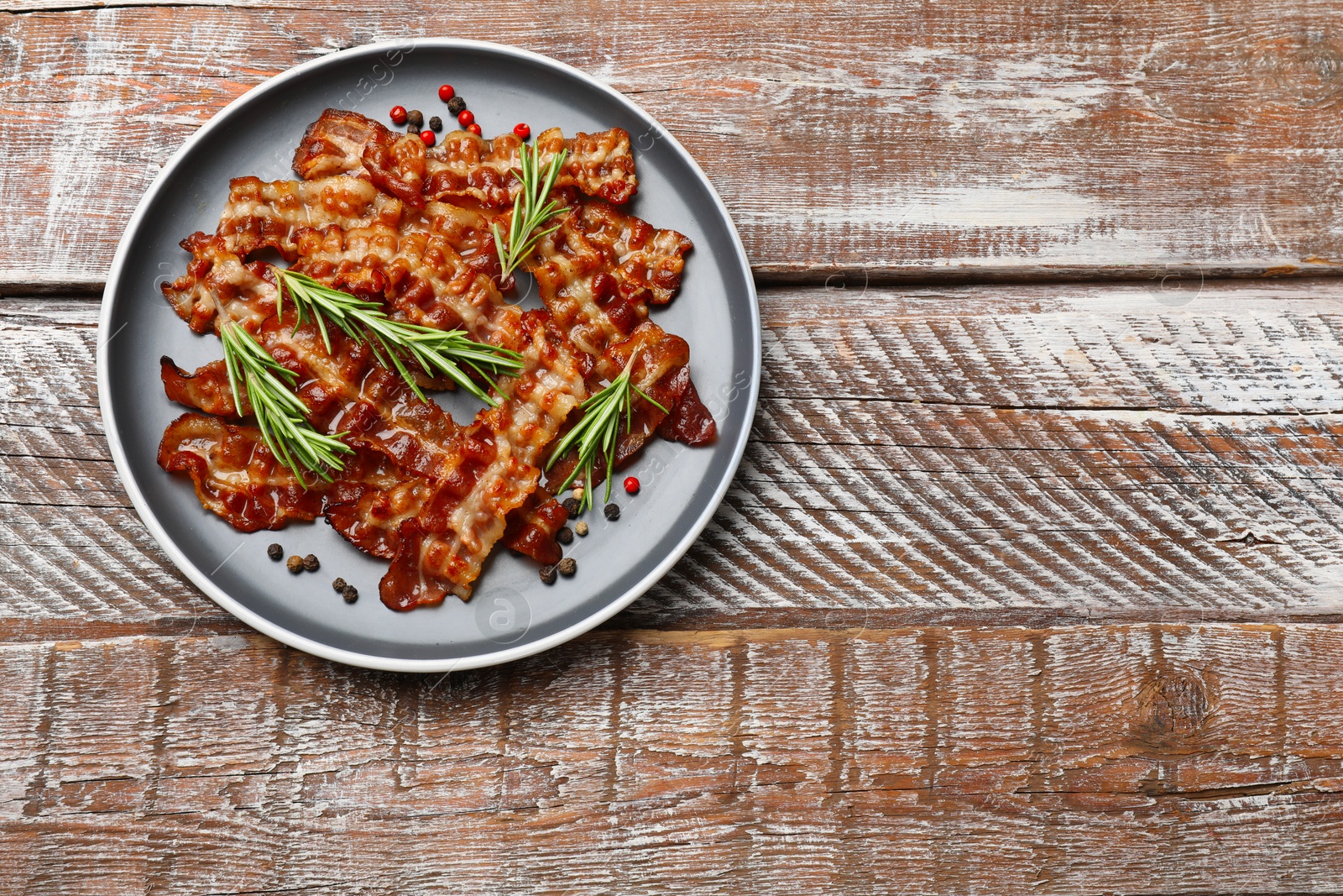 Photo of Slices of tasty fried bacon, peppercorns and rosemary on wooden table, top view. Space for text