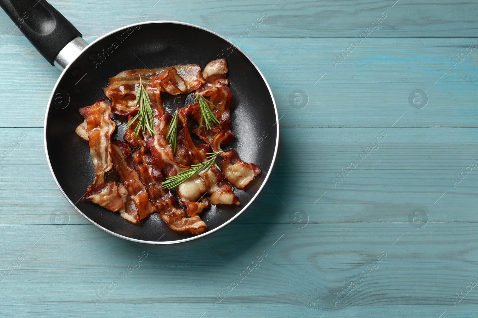 Photo of Delicious bacon slices in frying pan and rosemary on light blue wooden table, top view. Space for text