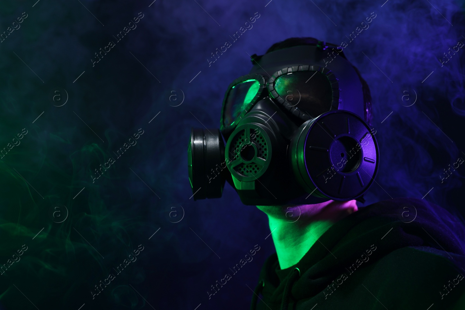 Photo of Man wearing gas mask in color lights and smoke on black background, low angle view. Space for text