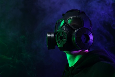 Photo of Man wearing gas mask in color lights and smoke on black background, low angle view. Space for text