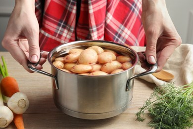 Woman holding pot with raw potatoes at wooden table, closeup